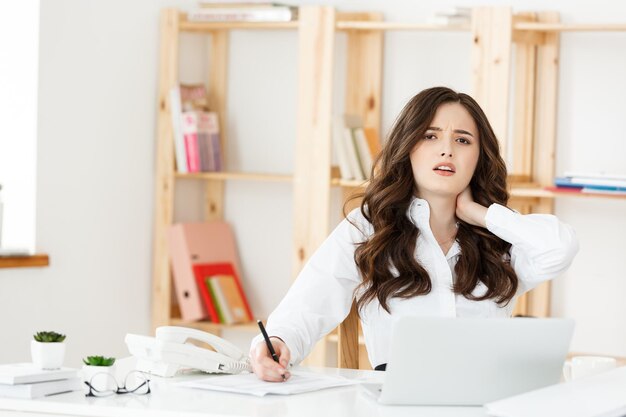 Giovane donna di affari stanca che soffre da molto tempo che si siede allo scrittorio del computer in ufficio