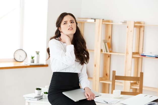 Giovane donna di affari stanca che soffre da molto tempo che si siede allo scrittorio del computer in ufficio