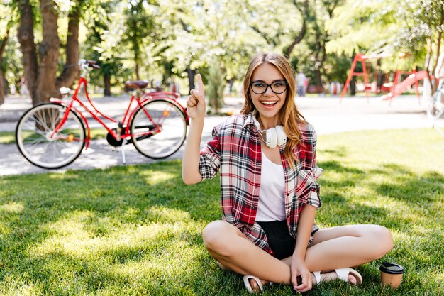 Giovane donna di buon umore seduta sull'erba con un sorriso sincero. Foto all'aperto di allegra ragazza caucasica agghiacciante nel parco.