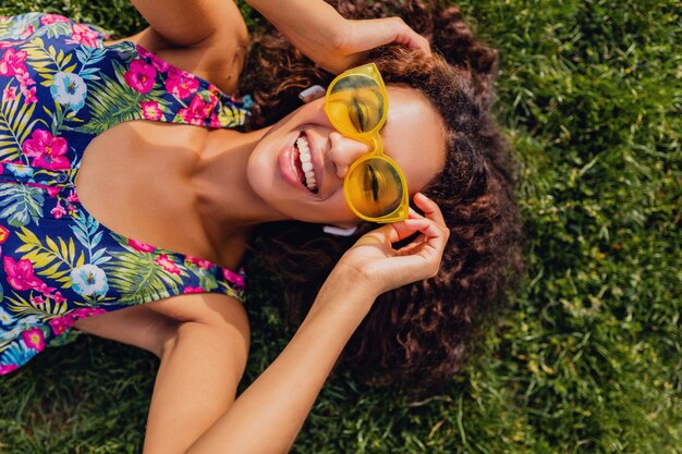 Giovane donna di colore alla moda che ascolta la musica sugli auricolari wireless divertendosi nel parco, stile di moda estiva, vestito colorato hipster, sdraiato sull'erba, vista dall'alto