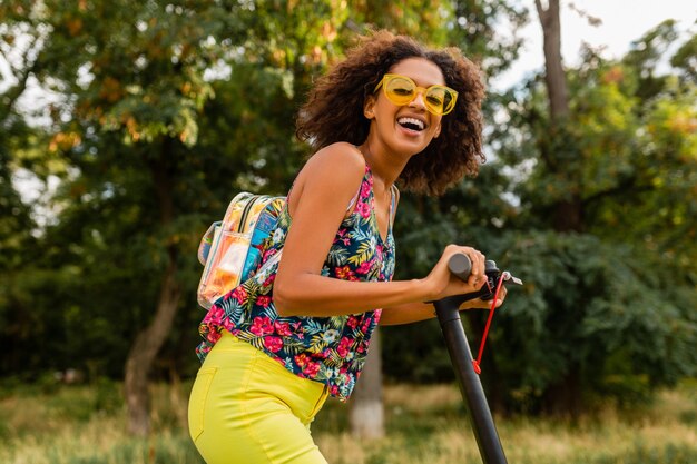 Giovane donna di colore alla moda che si diverte nel parco cavalcando scooter elettrico in stile moda estiva, vestito colorato hipster, indossando zaino e occhiali da sole gialli