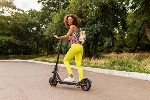 Giovane donna di colore alla moda che si diverte nel parco cavalcando scooter elettrico in stile moda estiva, vestito colorato hipster, indossando zaino e pantaloni gialli e occhiali da sole