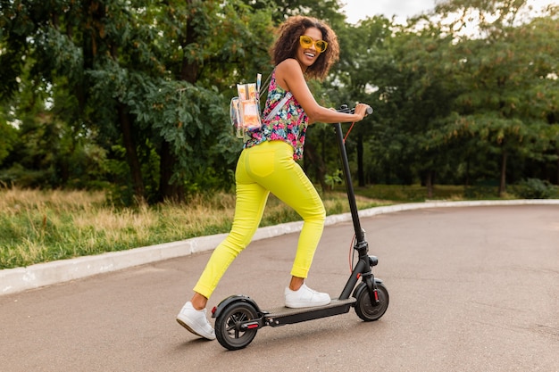 Giovane donna di colore alla moda che si diverte nel parco cavalcando scooter elettrico in stile moda estiva, vestito colorato hipster, indossando zaino e pantaloni gialli e occhiali da sole