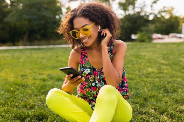 Giovane donna di colore sorridente elegante che utilizza smartphone che ascolta la musica sugli auricolari senza fili divertendosi nel parco