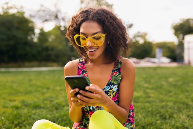 Giovane donna di colore sorridente elegante che utilizza smartphone che ascolta la musica sugli auricolari senza fili divertendosi nel parco