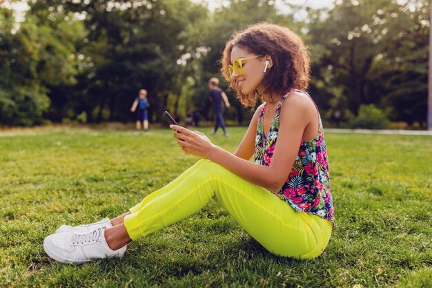 Giovane donna di colore sorridente elegante che utilizza smartphone che ascolta la musica sugli auricolari wireless divertendosi nel parco, stile colorato di moda estiva