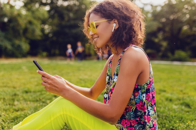 Giovane donna di colore sorridente elegante che utilizza smartphone che ascolta la musica sugli auricolari wireless divertendosi nel parco, stile colorato di moda estiva