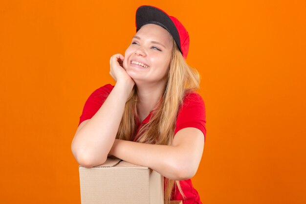 Giovane donna di consegna che indossa la maglietta polo rossa e cappello sorridente in attesa tenendo la mano sulla guancia mentre lo sostengono con un'altra mano incrociata guardando fiducioso e felice su sfondo arancione isolato