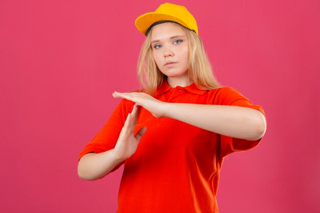 Giovane donna di consegna che indossa la maglietta polo rossa e cappuccio giallo cercando oberati di lavoro rendendo il gesto di time out con le mani su sfondo rosa isolato