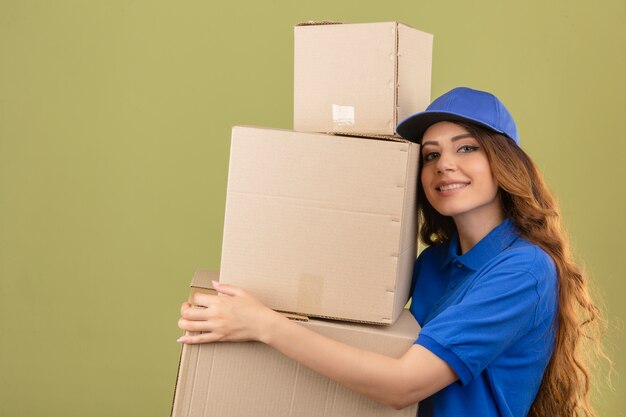 Giovane donna di consegna con capelli ricci che indossa la maglietta polo blu e cappuccio in piedi con una pila di scatole di cartone in mani sorridenti amichevoli su sfondo verde isolato