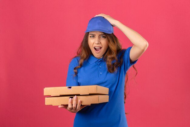 Giovane donna di consegna con i capelli ricci che indossa la maglietta polo blu e il cappuccio in piedi con scatole per pizza scioccato con la mano sulla testa per errore ricorda l'errore dimenticato il concetto di cattiva memoria su rosa isolato b