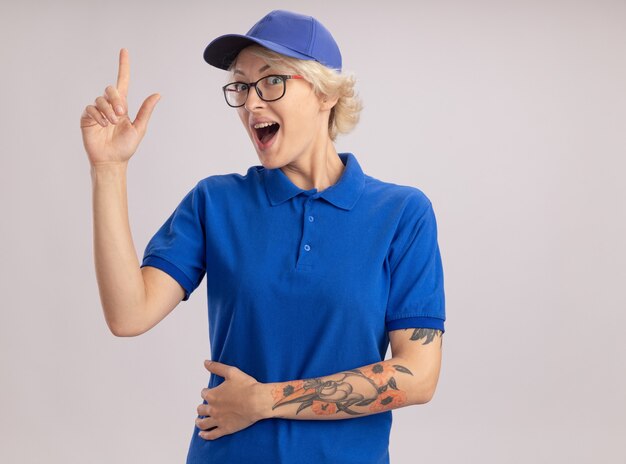 Giovane donna di consegna in uniforme blu e berretto felice e allegro che mostra il dito indice in piedi sopra il muro bianco