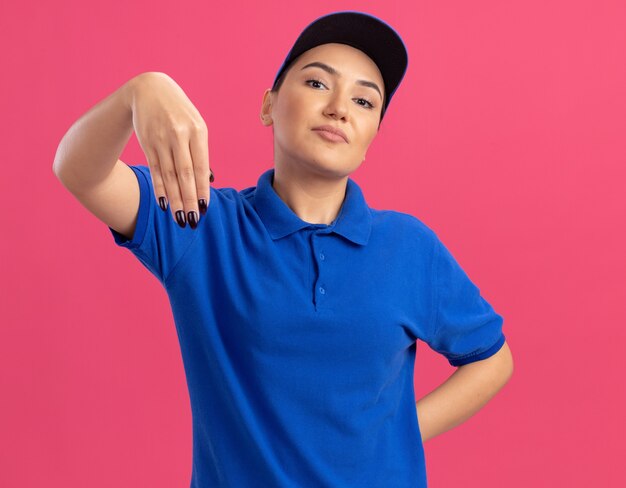 Giovane donna di consegna in uniforme blu e berretto guardando fiducioso gesticolando con le mani in piedi sopra la parete rosa