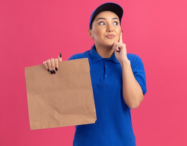 Giovane donna di consegna in uniforme blu e cappuccio che tiene il pacchetto di carta che osserva in su con il sorriso sul fronte che sta sopra la parete rosa