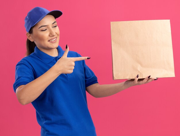 Giovane donna di consegna in uniforme blu e cappuccio che tiene il pacchetto di carta che punta con il dito indice sorridendo fiducioso in piedi sopra il muro rosa