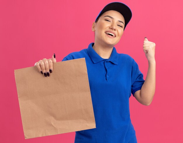 Giovane donna di consegna in uniforme blu e cappuccio che tiene il pacchetto di carta felice ed emozionato che alza il pugno che sorride allegramente in piedi sopra la parete rosa