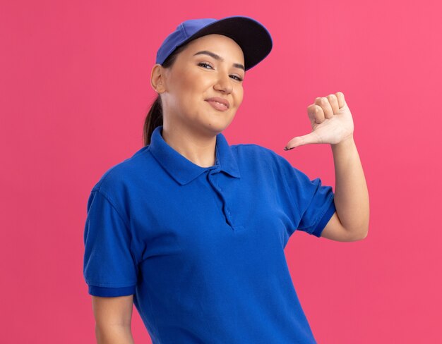 Giovane donna di consegna in uniforme blu e cappuccio felice e fiducioso che indica con il pollice a se stessa in piedi sopra la parete rosa