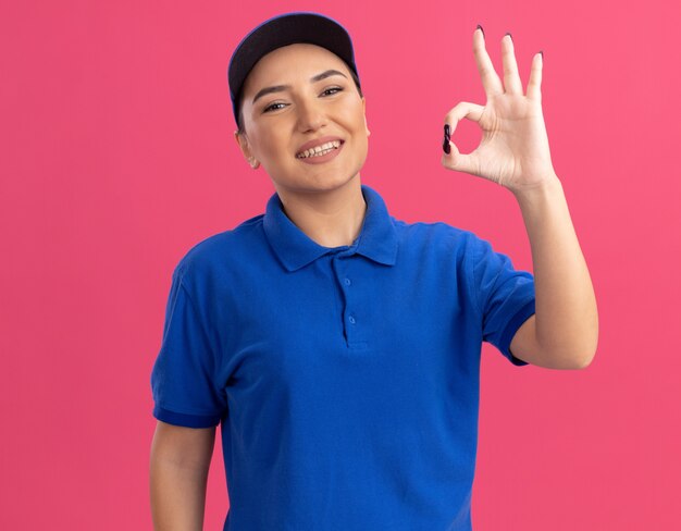 Giovane donna di consegna in uniforme blu e cappuccio guardando davanti sorridente fiducioso che mostra segno giusto in piedi sopra il muro rosa