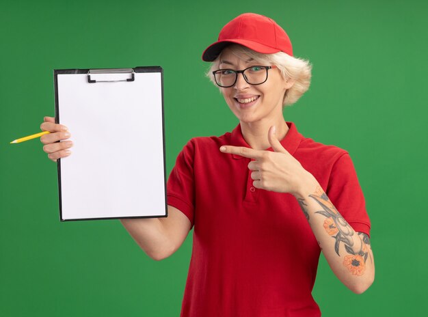 Giovane donna di consegna in uniforme rossa e berretto con gli occhiali che mostra appunti con pagine vuote e matita che punta con il dito indice negli appunti sorridendo allegramente in piedi sopra la parete verde