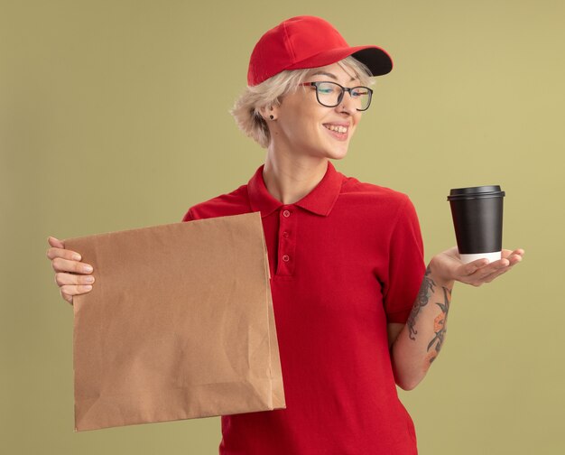 Giovane donna di consegna in uniforme rossa e berretto con gli occhiali che tiene il pacchetto di carta e il bicchiere di carta che sembra sorridente sicuro in piedi sopra la parete verde