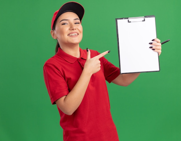 Giovane donna di consegna in uniforme rossa e cappuccio che tiene appunti con pagine vuote che punta con il dito indice negli appunti guardando davanti sorridendo allegramente in piedi sopra la parete verde