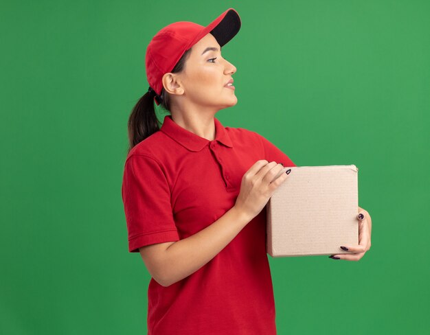 Giovane donna di consegna in uniforme rossa e cappuccio che tiene la scatola di cartone che guarda da parte con il sorriso sul viso in piedi sopra la parete verde