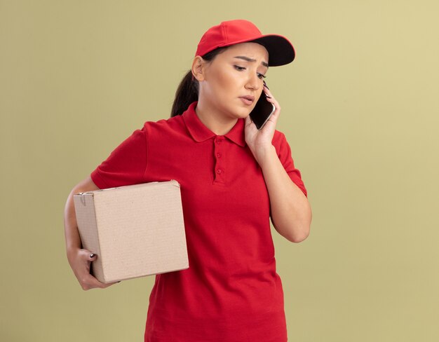 Giovane donna di consegna in uniforme rossa e cappuccio che tiene la scatola di cartone che sembra confusa mentre parla al telefono cellulare in piedi sopra la parete verde