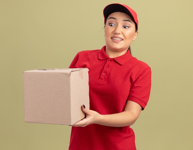 Giovane donna di consegna in uniforme rossa e cappuccio che tiene la scatola di cartone guardandolo felice e sorpreso in piedi sopra la parete verde