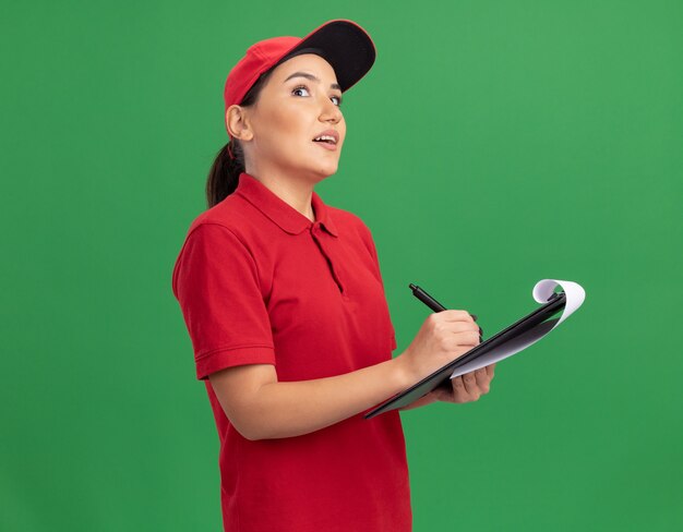 Giovane donna di consegna in uniforme rossa e cappuccio con appunti e matita che osserva in su con espressione pensierosa pensando in piedi sopra la parete verde