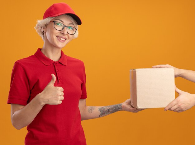 Giovane donna di consegna in uniforme rossa e cappuccio sorridente amichevole che mostra i pollici in su mentre riceve il pacchetto della scatola che sta sopra la parete arancione