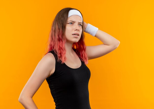Giovane donna di forma fisica in abbigliamento sportivo che osserva da parte confuso e molto ansioso in piedi sopra la parete arancione