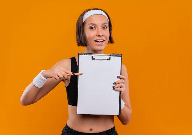 Giovane donna di forma fisica in abiti sportivi con fascia che mostra appunti con pagine vuote che punta con la penna ad esso sorridente in piedi amichevole sopra la parete arancione