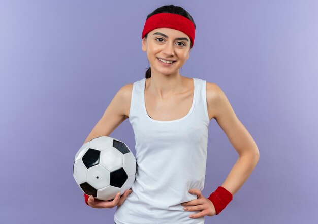 Giovane donna di forma fisica in abiti sportivi con fascia che tiene pallone da calcio sorridente fiducioso in piedi felice e positivo sopra la parete viola