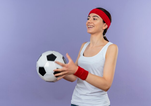 Giovane donna di forma fisica in abiti sportivi con fascia che tiene pallone da calcio sorridente fiducioso in piedi felice e positivo sopra la parete viola