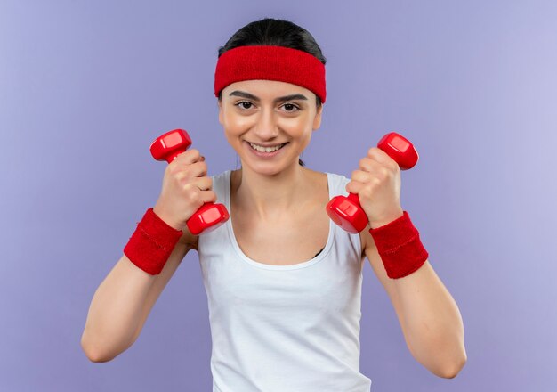 Giovane donna di forma fisica in abiti sportivi con fascia tenendo due manubri in mani alzate