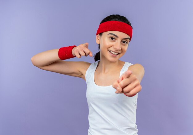Giovane donna di forma fisica in abiti sportivi con la fascia che punta con il dito indice alla fotocamera sorridendo allegramente in piedi sopra la parete viola