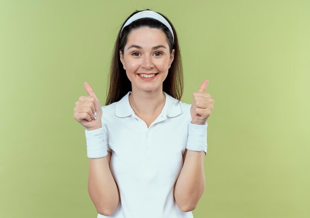 Giovane donna di forma fisica in fascia che sorride allegramente mostrando i pollici in su in piedi sopra la parete leggera