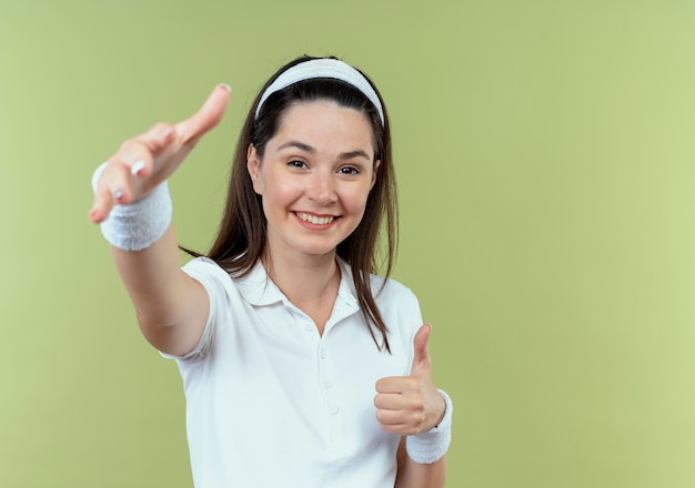 Giovane donna di forma fisica nella fascia felice e positiva che fa gesto di benvenuto con le mani in piedi su sfondo chiaro