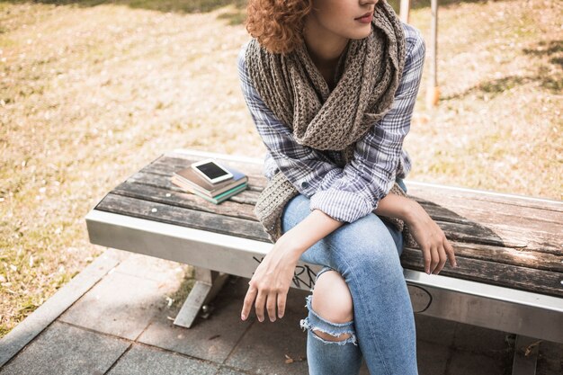 Giovane donna di redhead in sciarpa lavorata a maglia che si siede sul banco nel parco