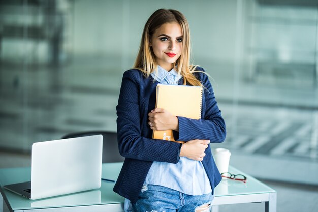 Giovane donna di successo in abiti casual che tengono notebook lavoro in piedi vicino alla scrivania bianca con il computer portatile in ufficio. Realizzazione del concetto di carriera aziendale.