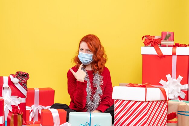 Giovane donna di vista frontale che si siede intorno ai regali di natale con la maschera sul colore giallo del nuovo anno del covid di emozione del virus