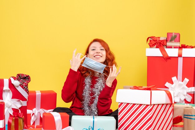 Giovane donna di vista frontale che si siede intorno ai regali di natale con la maschera sul virus giallo di colore di covid di emozione del nuovo anno