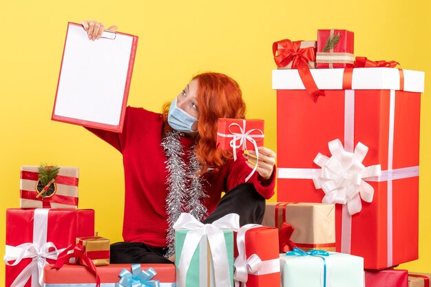 Giovane donna di vista frontale che si siede intorno ai regali di natale con la nota del file sul colore giallo covid del nuovo anno del virus dell'emozione