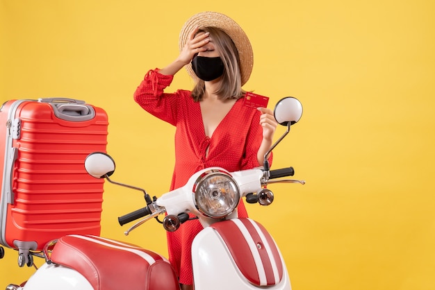 Giovane donna di vista frontale con la maschera nera che tiene la carta di credito che sta vicino alla valigia rossa del motorino