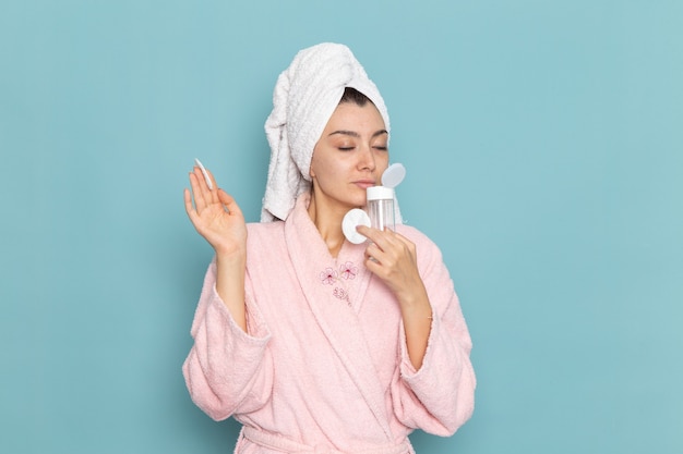 Giovane donna di vista frontale in accappatoio rosa che tiene lo struccante sulla parete blu pulizia bellezza acqua pulita doccia crema selfcare