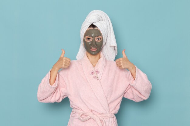 Giovane donna di vista frontale in accappatoio rosa dopo la doccia con la maschera scura sul viso sulla scrivania blu bellezza acqua crema selfcare doccia bagno