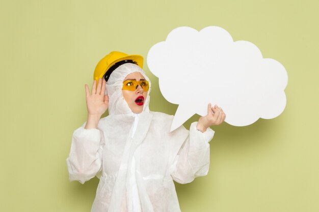 Giovane donna di vista frontale in vestito speciale bianco e casco protettivo giallo che tiene grande segno bianco sulla scienza uniforme della tuta spaziale verde