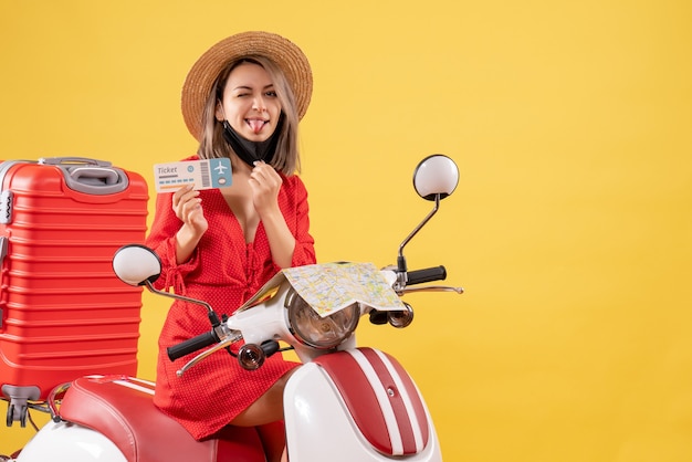 Giovane donna di vista frontale sul ciclomotore con la valigia rossa che sporge il biglietto della holding della lingua tongue