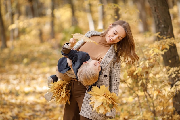 Giovane donna e ragazzino nella foresta di autunno. Donna che tiene suo figlio sulle mani. Ragazzo che indossa abiti di moda e tiene foglie gialle.