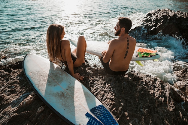 Giovane donna e uomo con tavole da surf seduti su roccia vicino al mare